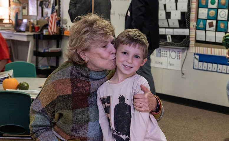 A woman kisses a young boy on the cheek in a classroom setting.